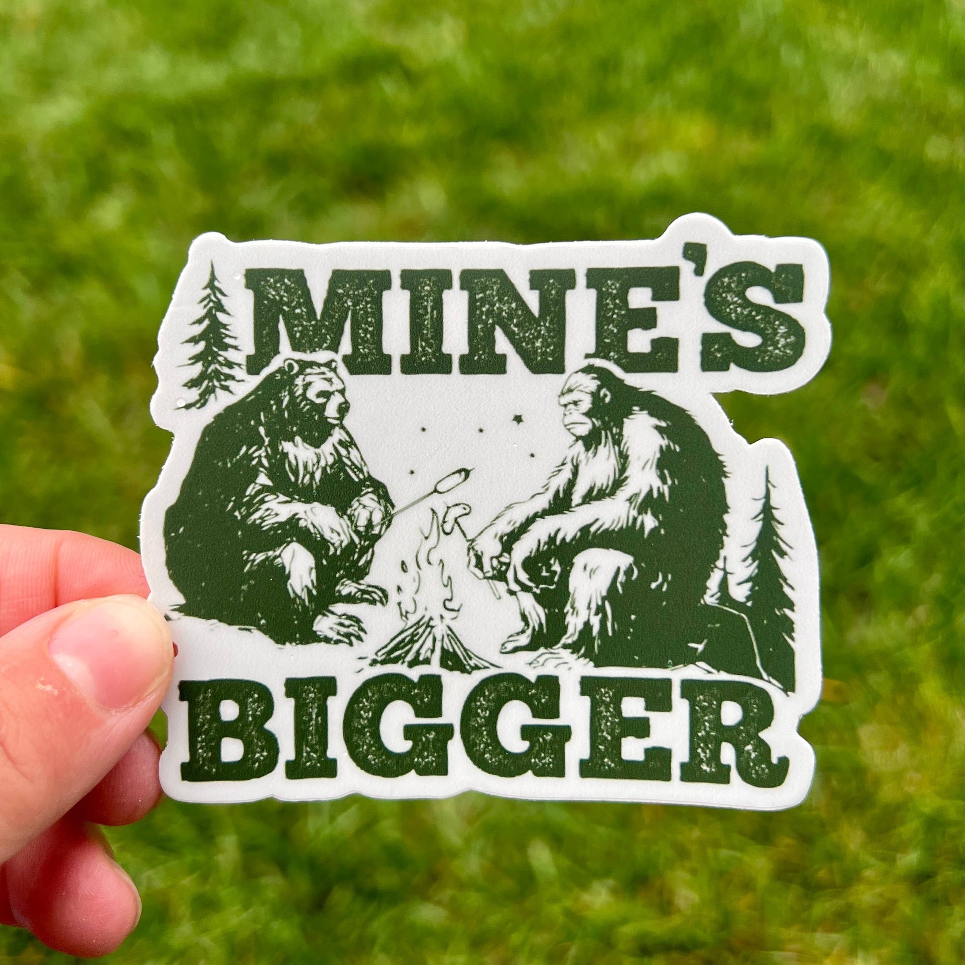 Sticker with bear and Sasquatch design held by a hand against a grassy background